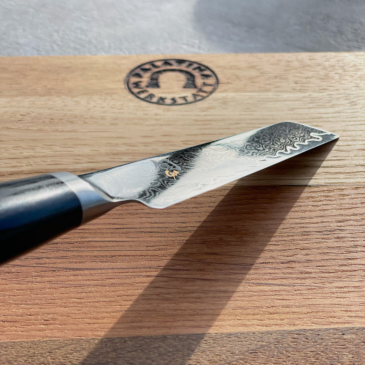 Eine Nahaufnahme eines hochwertigen Kai Shun Premier Minamo TMM-0700 Büromessers mit einer 9 cm langen Damaststahlklinge, platziert auf einem 30x22 cm großen Küchenbrett aus Eiche. Das Schneidebrett weist ein eingraviertes kreisförmiges Logo auf. Der dunkle Griff des Messers kontrastiert mit seiner glänzenden, aufwendig gemusterten Klinge.