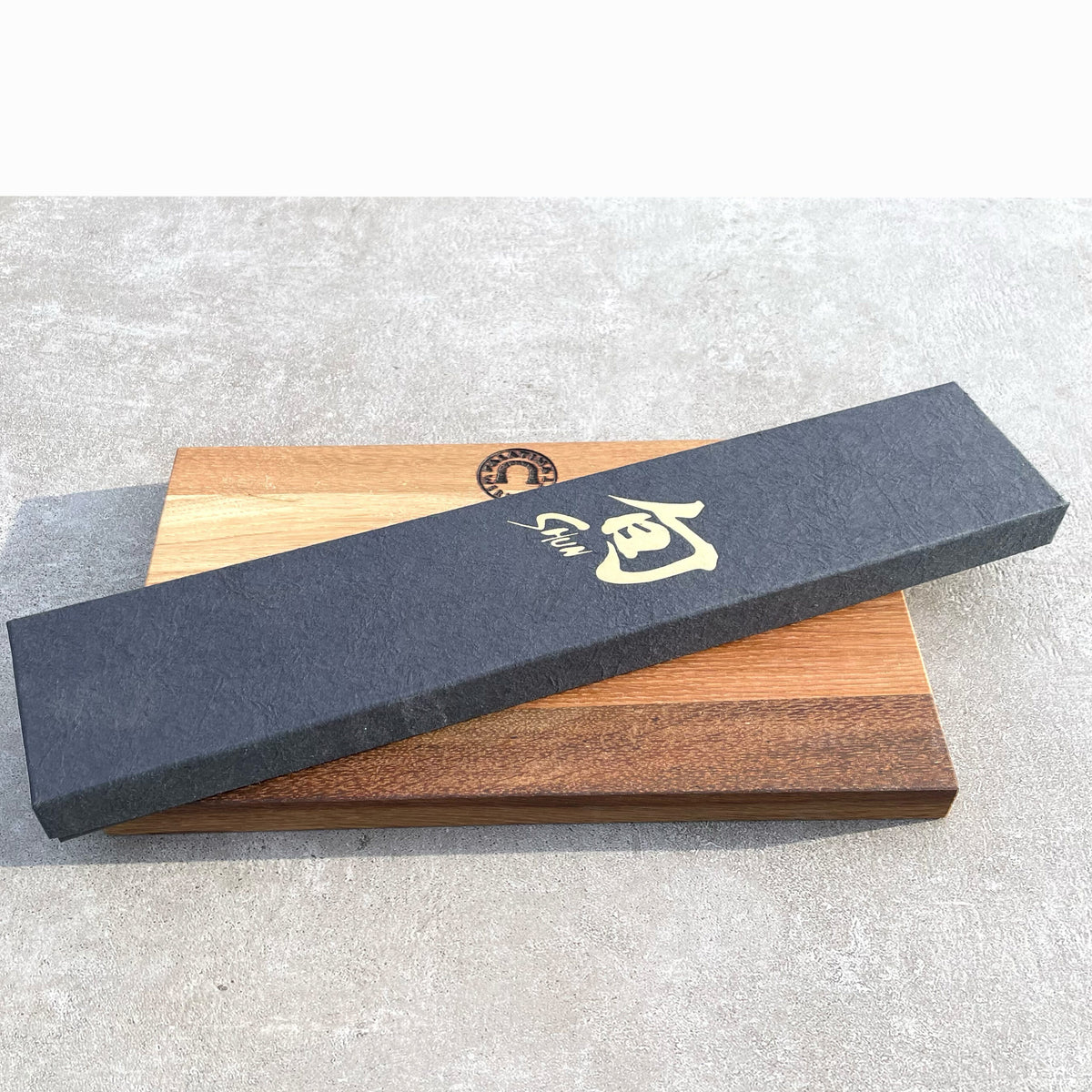 Ein ultrascharfes Santoku Kai Shun DM-0702 mit 18 cm Klinge, versehen mit einem rechteckigen schwarzen Kästchen mit goldener Schrift, liegt auf einem handgefertigten hellbraunen Fassholzbrett (30x18 cm). Der Hintergrund ist eine strukturierte graue Oberfläche.