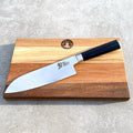 Das Kai Shun DM-0702 ultrascharfes Santoku mit einer 18 cm langen Klinge liegt auf einem handgefertigten Fassholzbrett mit hellen und dunklen Streifen. Der schwarze Griff des Messers wird von japanischen Inschriften auf der Klinge ergänzt. Das Schneidebrett zeigt ein rundes Logo in der Mitte nahe dem oberen Rand, alles auf einer Betonfläche arrangiert.
