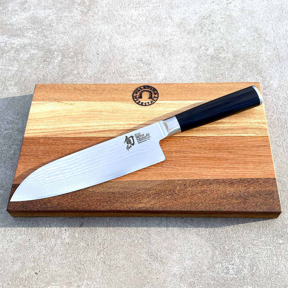 Das Kai Shun DM-0702 ultrascharfes Santoku mit einer 18 cm langen Klinge liegt auf einem handgefertigten Fassholzbrett mit hellen und dunklen Streifen. Der schwarze Griff des Messers wird von japanischen Inschriften auf der Klinge ergänzt. Das Schneidebrett zeigt ein rundes Logo in der Mitte nahe dem oberen Rand, alles auf einer Betonfläche arrangiert.