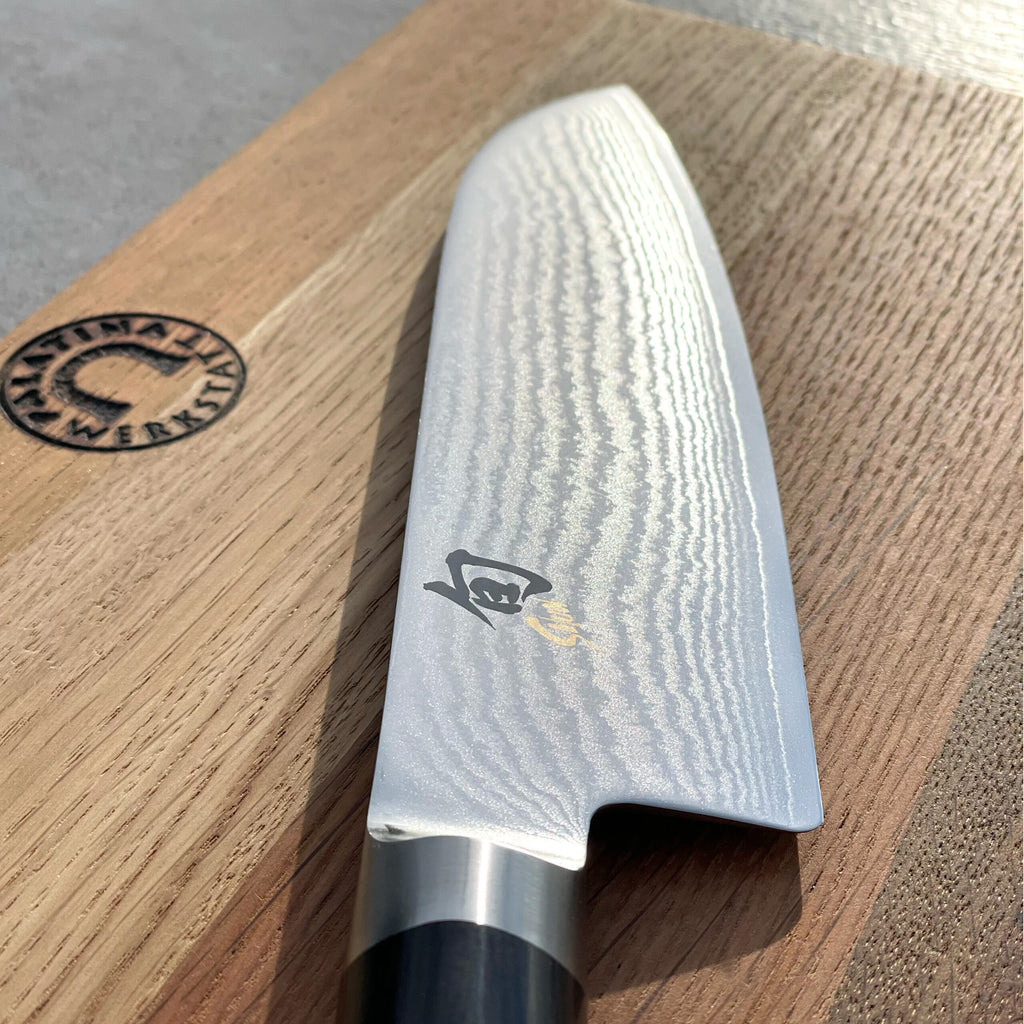 Eine Nahaufnahme des polierten, ultrascharfen Santoku Kai Shun DM-0702 mit einer 18 cm langen Klinge aus Damaszenerstahl, das auf einem handgefertigten 30 x 18 cm großen Fassholzbrett ruht. Das Messer hat einen dunklen Griff mit einem kleinen Emblem nahe der Klingenbasis. Die Holzmaserung ist sichtbar und die Beleuchtung hebt die Textur hervor.