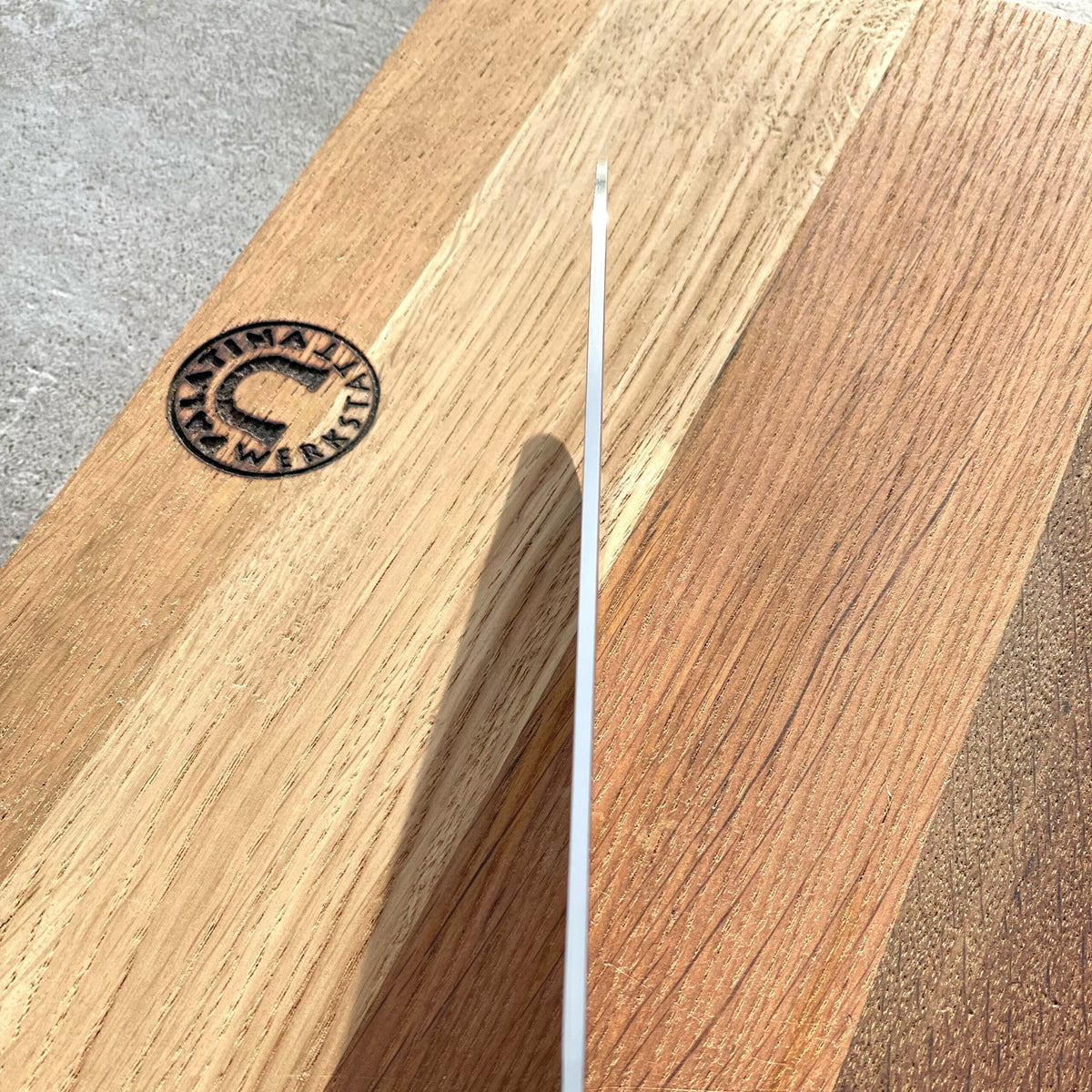 Ein dünnes Objekt, möglicherweise das ultrascharfe Santoku Kai Shun DM-0702 mit 18 cm Klinge, wird von der Seite auf einer Holzoberfläche fotografiert, die mehrere Farbtöne aufweist. Die Sonne wirft einen Schatten auf dieses Objekt. Die Oberfläche ähnelt der ausgeprägten Textur und dem kreisförmigen Logo des handgefertigten Fassholzbretts (30x18 cm).