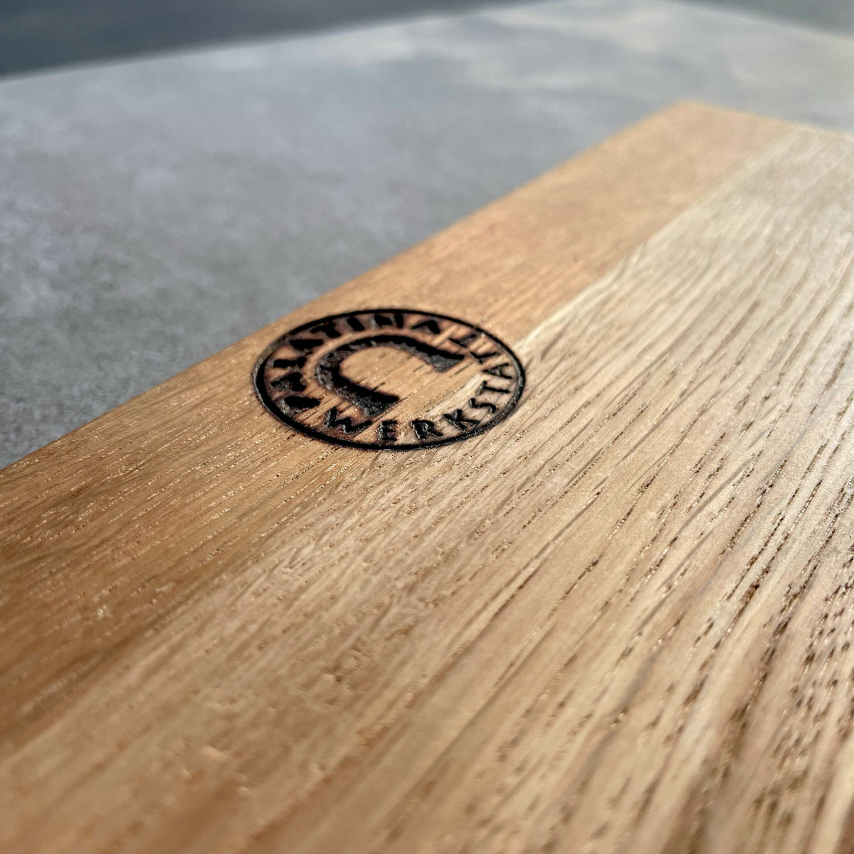 Eine Nahaufnahme einer Holzoberfläche mit einem eingebranntem kreisförmigen Logo. Das Logo zeigt ein Baumdesign mit den Worten „Nashville Custom Woodworking“, die es umgeben. Das Foto hebt die Textur und Maserung des dazugehörigen handgefertigten Fassholzbretts hervor, das 30 x 18 cm misst und mit dem ultrascharfen Santokumesser Kai Shun DM-0702 mit einer 18 cm langen Klinge gepaart ist.