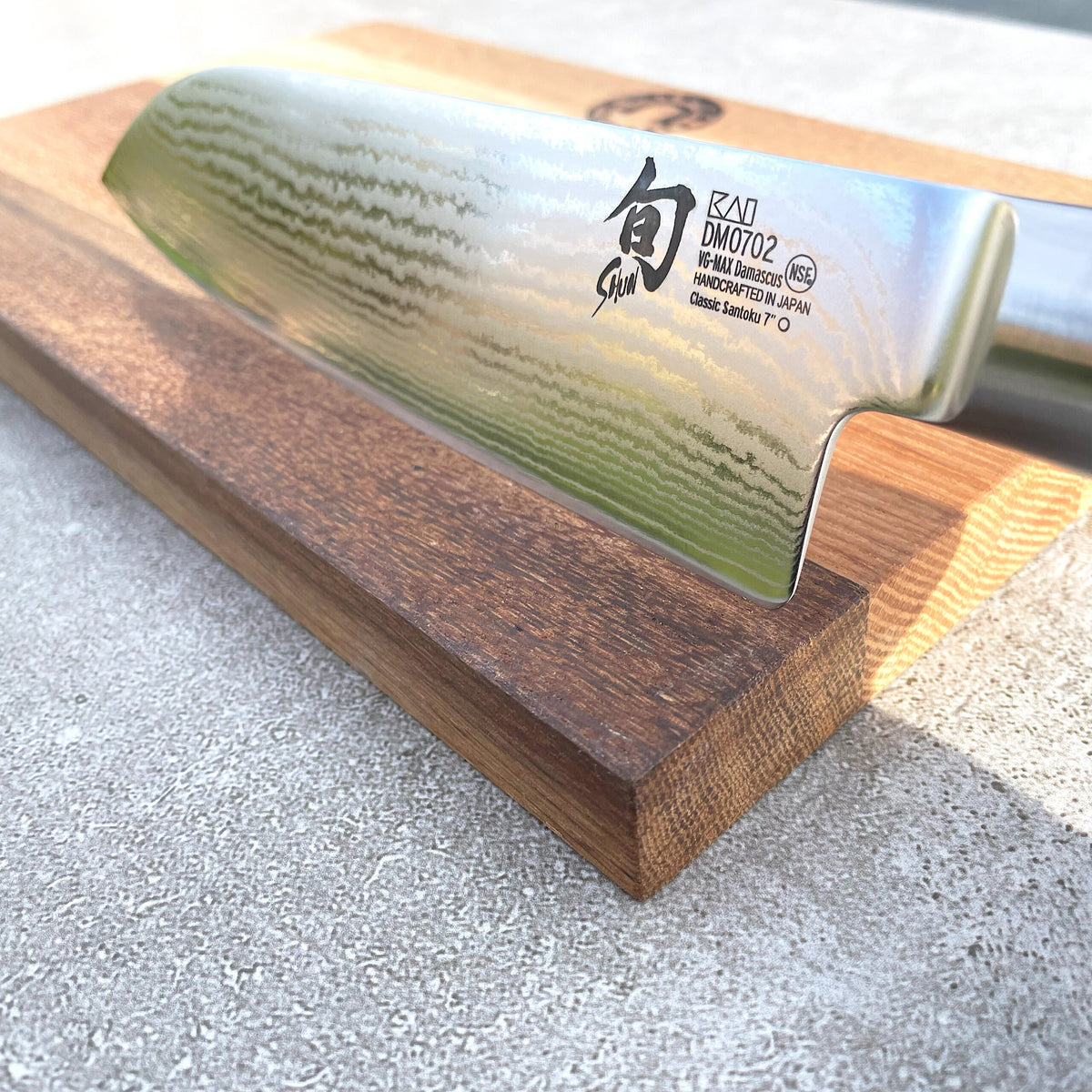 Nahaufnahme eines ultrascharfen japanischen Santokumessers Kai Shun DM-0702 mit einer 18 cm langen, wellenförmigen Damaszenerklinge mit schwarzen Kanji-Zeichen und englischen Angaben. Das Messer liegt auf einem handgefertigten Fassholzbrett mit den Maßen 30x18 cm, das auf einer Betonoberfläche steht.