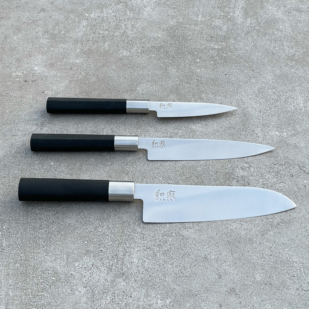In einem optisch ansprechenden Arrangement auf einer Betonfläche werden drei Messer aus dem 4-teiligen Geschenkset von KAI Weihnachtsangebot mit Wasabi Black-Griffen ausgestellt. Die Messer sind unterschiedlich groß, wobei das kleinste oben und das größte unten positioniert ist. Auf der Klinge sind jeweils japanische Schriftzeichen eingraviert.