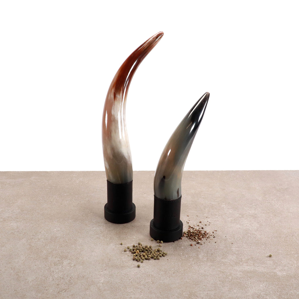 Zwei exklusive Pfeffermühlen aus echtem Horn von Forge de Laguiole – eine braun und weiß, die andere grau und schwarz – stehen aufrecht auf einer neutral gefärbten Oberfläche. Kleine Häufchen grüner und schwarzer Pfefferkörner sind am Fuß jeder handgefertigten Pfeffermühle verstreut. Der Hintergrund ist weiß und minimalistisch.
