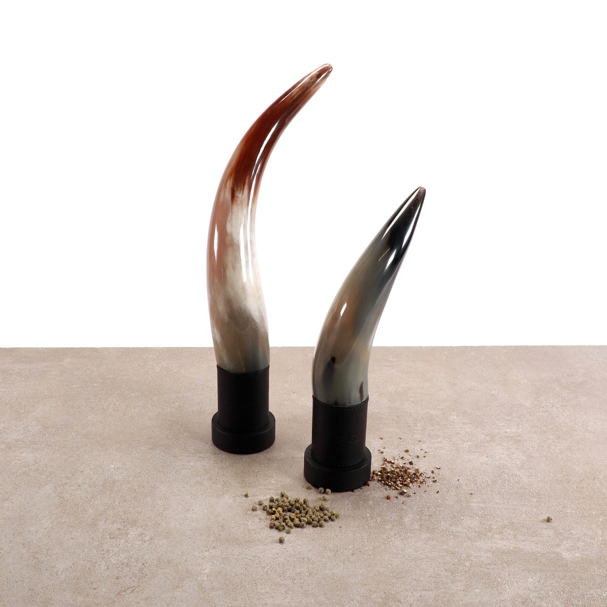 Zwei exklusive Pfeffermühlen aus echtem Horn von Forge de Laguiole – eine braun und weiß, die andere grau und schwarz – stehen aufrecht auf einer neutral gefärbten Oberfläche. Kleine Häufchen grüner und schwarzer Pfefferkörner sind am Fuß jeder handgefertigten Pfeffermühle verstreut. Der Hintergrund ist weiß und minimalistisch.