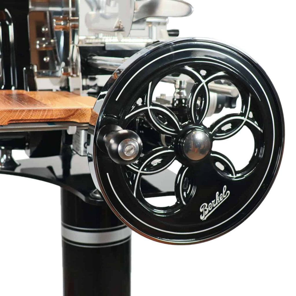 Nahaufnahme der schwarzen Berkel Volano B2 Schwungradmaschine im Vintage-Stil. Die Aufschnittmaschine zeichnet sich durch aufwendige Metallarbeiten, eine große Handkurbel und eine einzigartige hölzerne Schneidefläche aus. Das Berkel-Logo ist deutlich auf dem Griff zu sehen. Diese Schwungradmaschine zeigt mit ihrer atemberaubenden Handwerkskunst eine Mischung aus modernen und klassischen Designelementen.