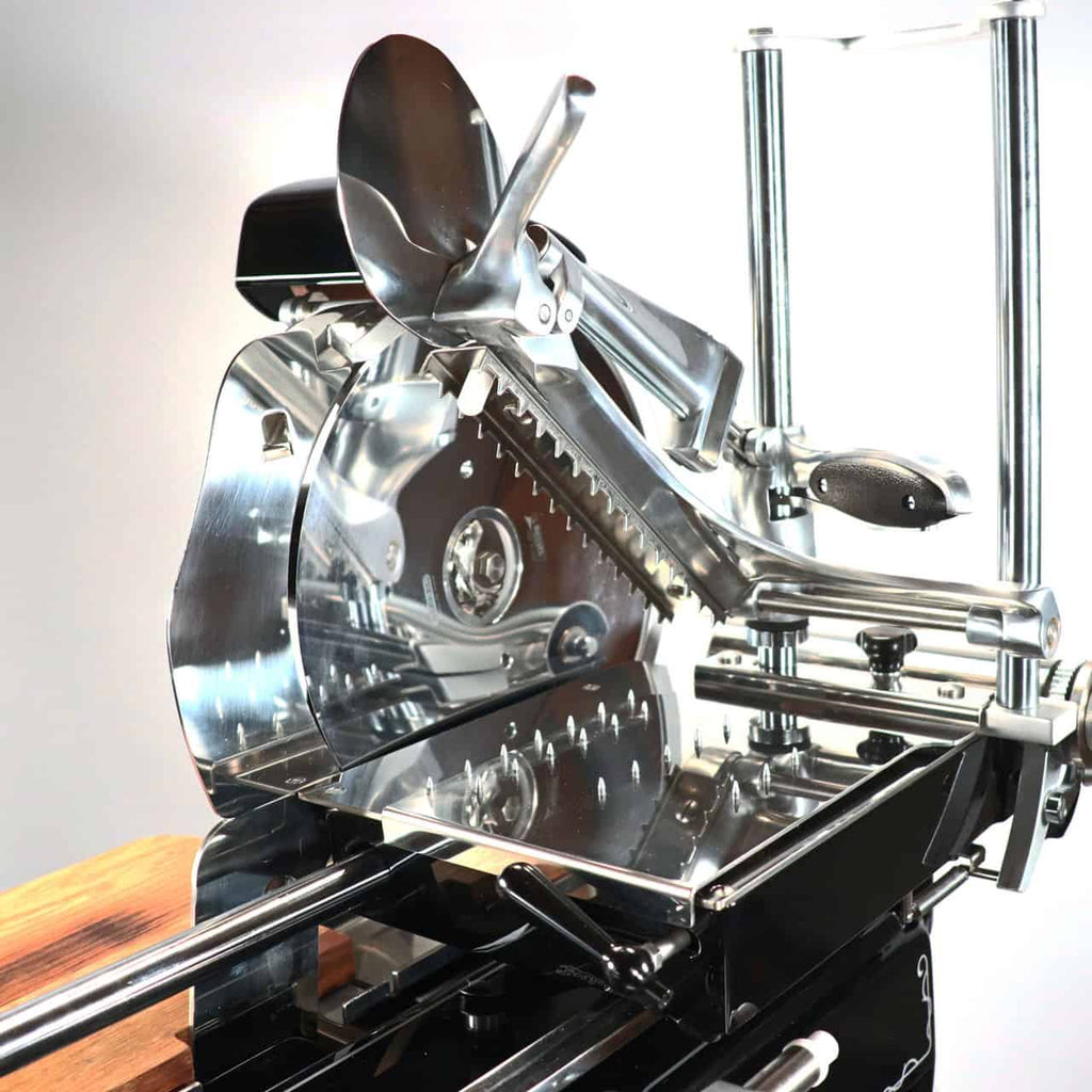 Die Berkel Volano B2 Schwungradmaschine in schwarz, ein atemberaubender manueller Allesschneider mit glänzender, metallischer Oberfläche und rotierender Klinge, steht auf einem schlichten Sockel. Er verfügt über einen großen Griff für die manuelle Bedienung und einen dazugehörigen Ständer mit einzigartigem Holzblock. Der Hintergrund ist in einer neutralen, hellen Farbe gehalten und betont das saubere und polierte Erscheinungsbild des Allesschneiders.