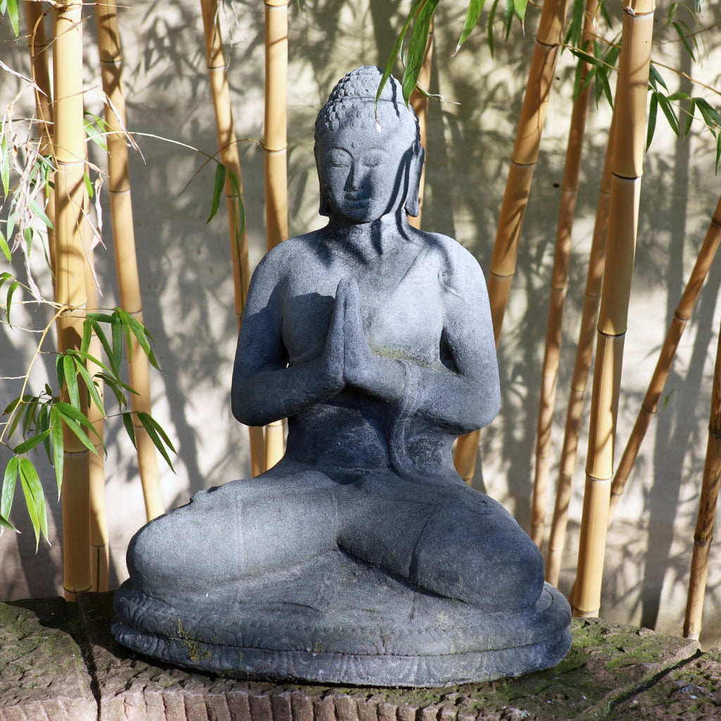 Eine Buddhastatue aus Stein sitzt in meditativer Pose mit den Händen in Gebetshaltung, umgeben von hohen Bambusstäben. Sonnenlicht fällt durch die Blätter und wirft Schatten auf die ruhige Figur und den Boden darunter. Diese sitzende Buddhafigur aus Lavastein 60 cm – Eine Quelle der inneren Ruhe strahlt Ruhe inmitten der Gelassenheit der Natur aus.