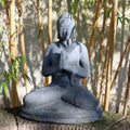 Eine Buddhastatue aus Stein sitzt in meditativer Pose mit den Händen in Gebetshaltung, umgeben von hohen Bambusstäben. Sonnenlicht fällt durch die Blätter und wirft Schatten auf die ruhige Figur und den Boden darunter. Diese sitzende Buddhafigur aus Lavastein 60 cm – Eine Quelle der inneren Ruhe strahlt Ruhe inmitten der Gelassenheit der Natur aus.