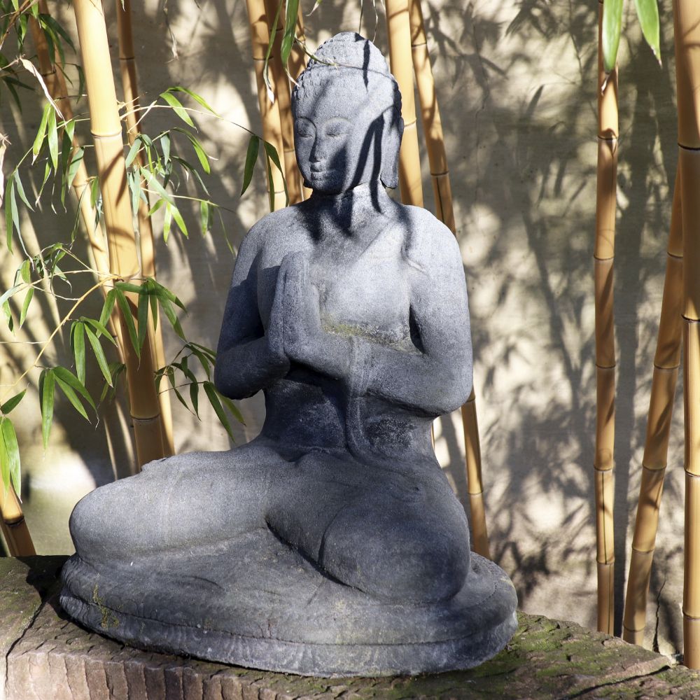 Eine Steinstatue eines sitzenden Buddhas in meditativer Pose, die Hände in Gebetshaltung, umgeben von Bambuspflanzen. Die sitzende Buddha-Figur aus Lavastein 60 cm – Eine Quelle der inneren Ruhe – Für Haus & Garten steht auf einer Holzoberfläche mit Schatten aus Bambusblättern, die die Figur bedecken und ein Gefühl innerer Ruhe hervorrufen.