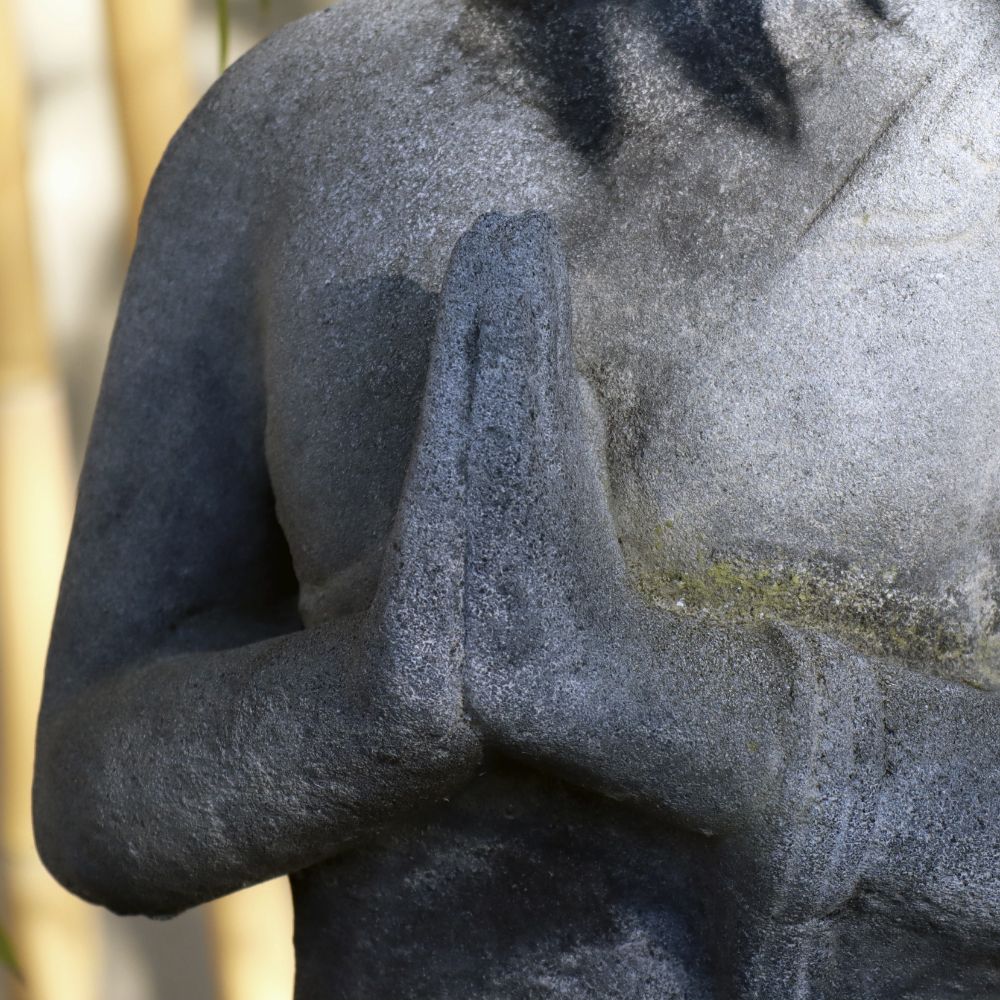 Eine Nahaufnahme einer verwitterten 60 cm großen Sitzenden Buddha-Figur aus Lavastein, die den Oberkörper und die Hände einer Person in Gebetshaltung zeigt. Die Textur des Steins zeigt Alterserscheinungen mit einigen Verfärbungen, was der Statue ein Aussehen verleiht, das innere Ruhe ausstrahlt. Der Hintergrund ist wunderschön verschwommen.