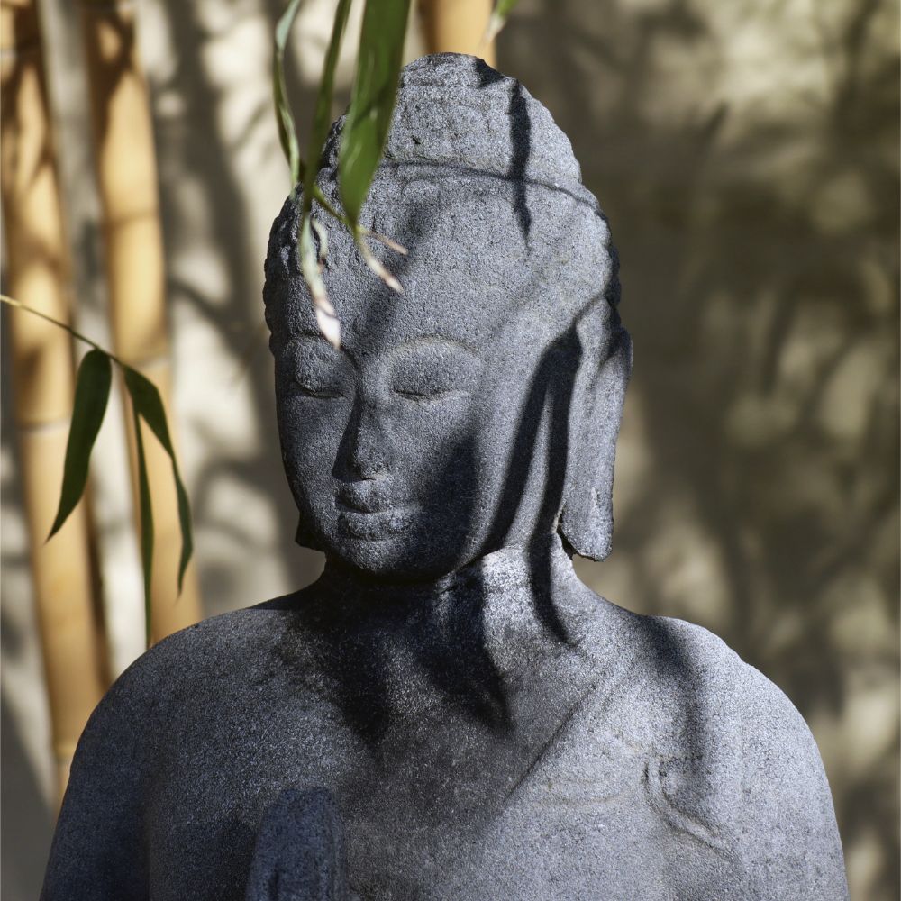 Eine Buddha-Statue aus Lavastein, die mit heiterem Gesichtsausdruck sitzt und teilweise von Bambusblättern beschattet wird, vor einem verschwommenen Hintergrund. Die geschlossenen Augen der Figur vermitteln ein Gefühl innerer Ruhe und Kontemplation. Das detailreiche Gewand fällt elegant über eine Schulter und strahlt Gelassenheit aus. Dies ist die „Sitzende Buddha-Figur aus Lavastein 60 cm – Eine Quelle der inneren Ruhe – Für Haus & Garten“.
