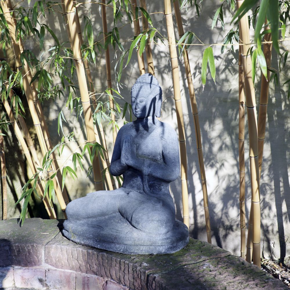 Eine sitzende Buddhafigur aus Lavastein (60 cm), eine Quelle der inneren Ruhe, steht auf einer Ziegelfläche mit grünen Bambuspflanzen und einer beigen Wand im Hintergrund. Sonnenlicht fällt durch den Bambus und erzeugt Schatten auf der Figur und der Wand. Der Buddha hält einen Gegenstand auf seinem Schoß, der innere Ruhe ausstrahlt.
