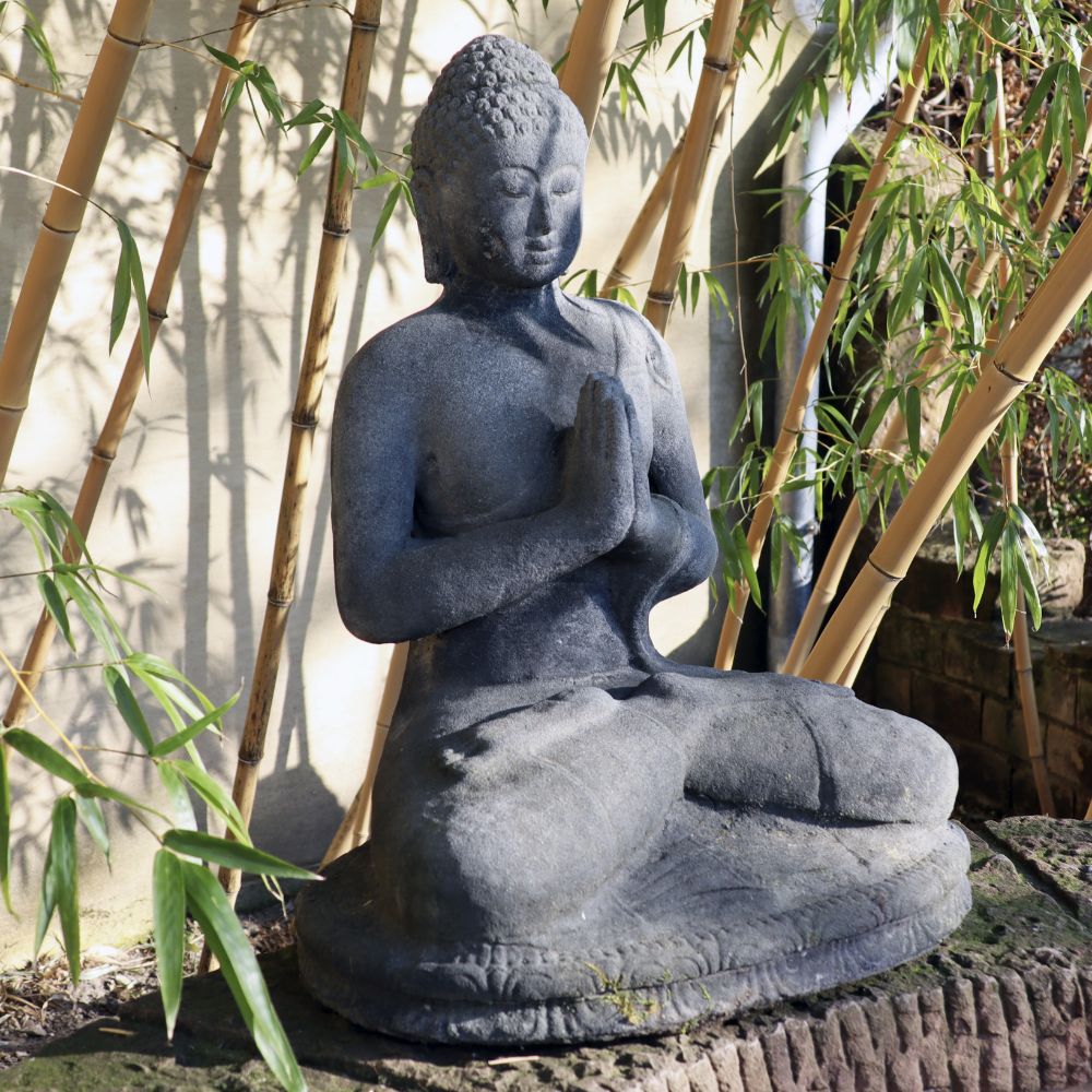 Eine sitzende Buddhafigur aus Lavastein (60 cm) befindet sich in einer meditativen Pose mit vor dem Herzen gefalteten Händen. Die Statue steht im Freien, ist von Bambuspflanzen umgeben und strahlt innere Ruhe aus.