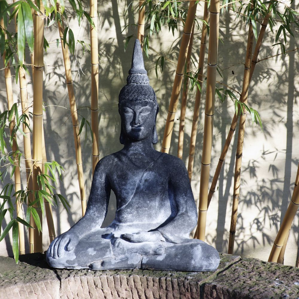 Die „Sitzende Buddha Figur aus Lavastein 75 cm – Eine Quelle der inneren Ruhe – Für Haus & Garten“ sitzt friedlich in einer meditativen Pose vor einem Hintergrund aus hohen Bambuspflanzen. Die aus Lavastein gefertigte Statue strahlt mit ihrem detailreichen Kopfschmuck und den heiteren, geschlossenen Augen, die teilweise vom Sonnenlicht beschattet werden, das durch die Blätter fällt, innere Ruhe aus.