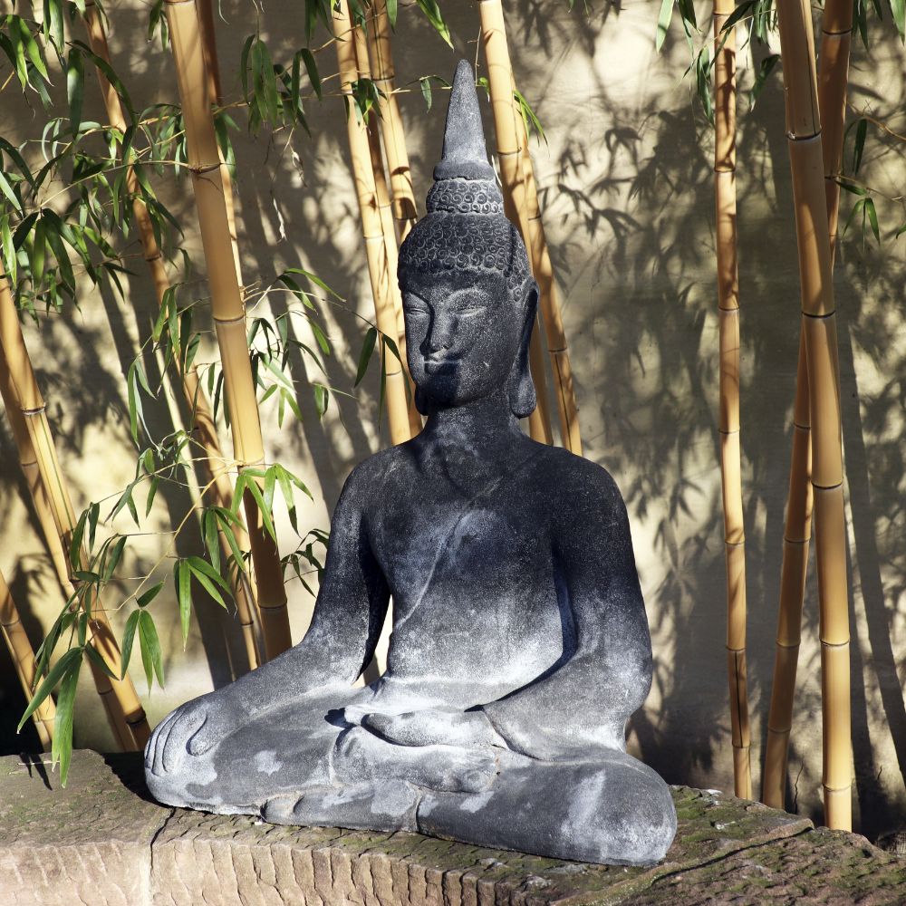 Eine ruhige, 75 cm große sitzende Buddhafigur aus Lavastein, eine Quelle der inneren Ruhe für Haus und Garten, sitzt in meditativer Pose mit gekreuzten Beinen und den Händen im Schoß. Umgeben von hohen Bambusstäben wirft sie komplizierte Schatten auf die Wand dahinter. Das umgebende Sonnenlicht verstärkt die friedliche Szene und verkörpert wahre innere Ruhe.