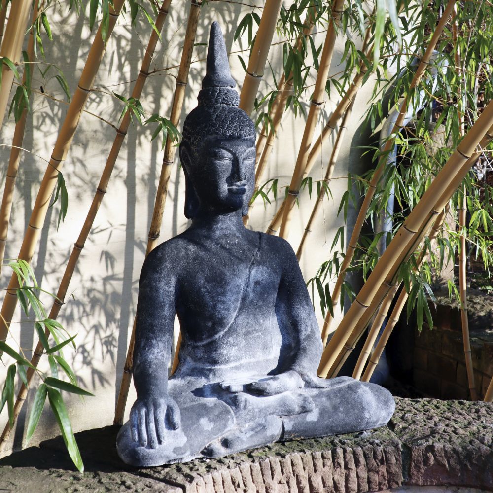 Eine sitzende Buddhafigur aus Lavastein 75cm - Eine Quelle der inneren Ruhe - Für Haus & Garten steht auf einer gemauerten Plattform. Die aus Lavastein gefertigte Statue steht vor einem Hintergrund aus Bambuspflanzen. Sonnenlicht wirft Schatten und verleiht der ruhigen und ruhigen Szene Tiefe, die inneren Frieden hervorruft.