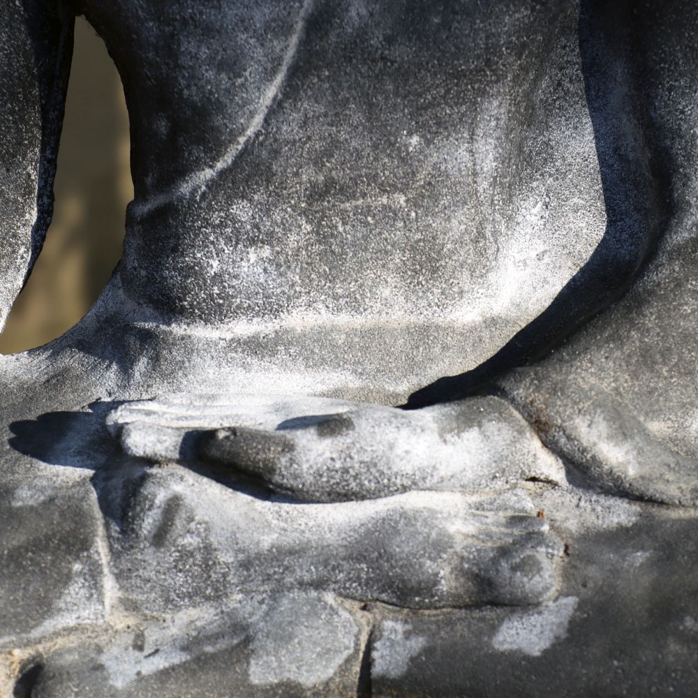 Eine Nahaufnahme der Sitzenden Buddha Figur aus Lavastein 75 cm – Eine Quelle der inneren Ruhe – Für Haus & Garten konzentriert sich auf den Oberkörper und die Hände in einer meditativen Haltung, wobei die rechte Handfläche auf der linken Handfläche ruht. Sonnenlicht wirft Schatten, betont die Textur und die abgenutzten Details der Statue und verkörpert eine Aura innerer Ruhe.