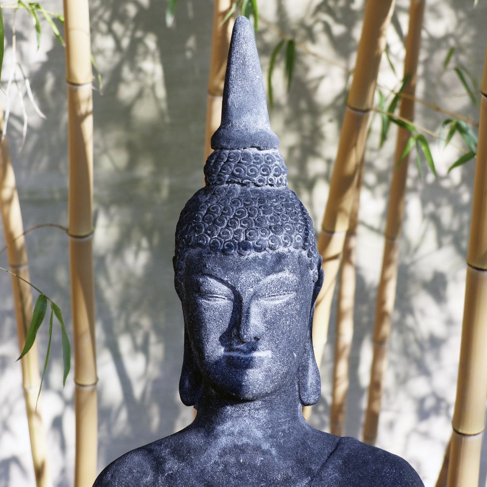 Eine Nahaufnahme zeigt die schwarze Steinstatue eines sitzenden Buddha, die aus Lavastein gefertigt und vom Sonnenlicht beleuchtet wird. Die Statue mit dem Namen „Sitzende Buddha Figur aus Lavastein 75 cm – Eine Quelle der inneren Ruhe“ steht im Freien, umgeben von hohen Bambusstäben und Blättern, die einen natürlichen und ruhigen Hintergrund bilden. Der ruhige und meditative Ausdruck des Buddha vermittelt auf wunderschöne Weise innere Ruhe.