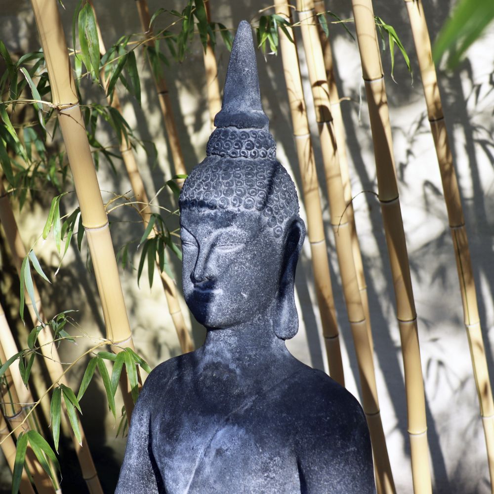 Eine ruhige Buddhastatue aus dunklem Stein, umgeben von hohen Bambusstämmen, durchflutet von sanftem Sonnenlicht. Die 75 cm große sitzende Buddha-Figur aus Lavastein – Eine Quelle der inneren Ruhe – für Haus & Garten hat eine spitze Ushnisha auf dem Kopf und zeigt einen ruhigen und meditativen Gesichtsausdruck, der innere Ruhe verkörpert.