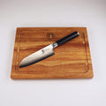 Das Kai Shun Classic Angebotsset DM-0727 verfügt über ein ultrascharfes 14 cm Santokumesser mit gemusterter Klinge und schwarzem Griff, das diagonal auf dem mitgelieferten massiven 30x18 cm Schneidebrett aus Eiche ruht.