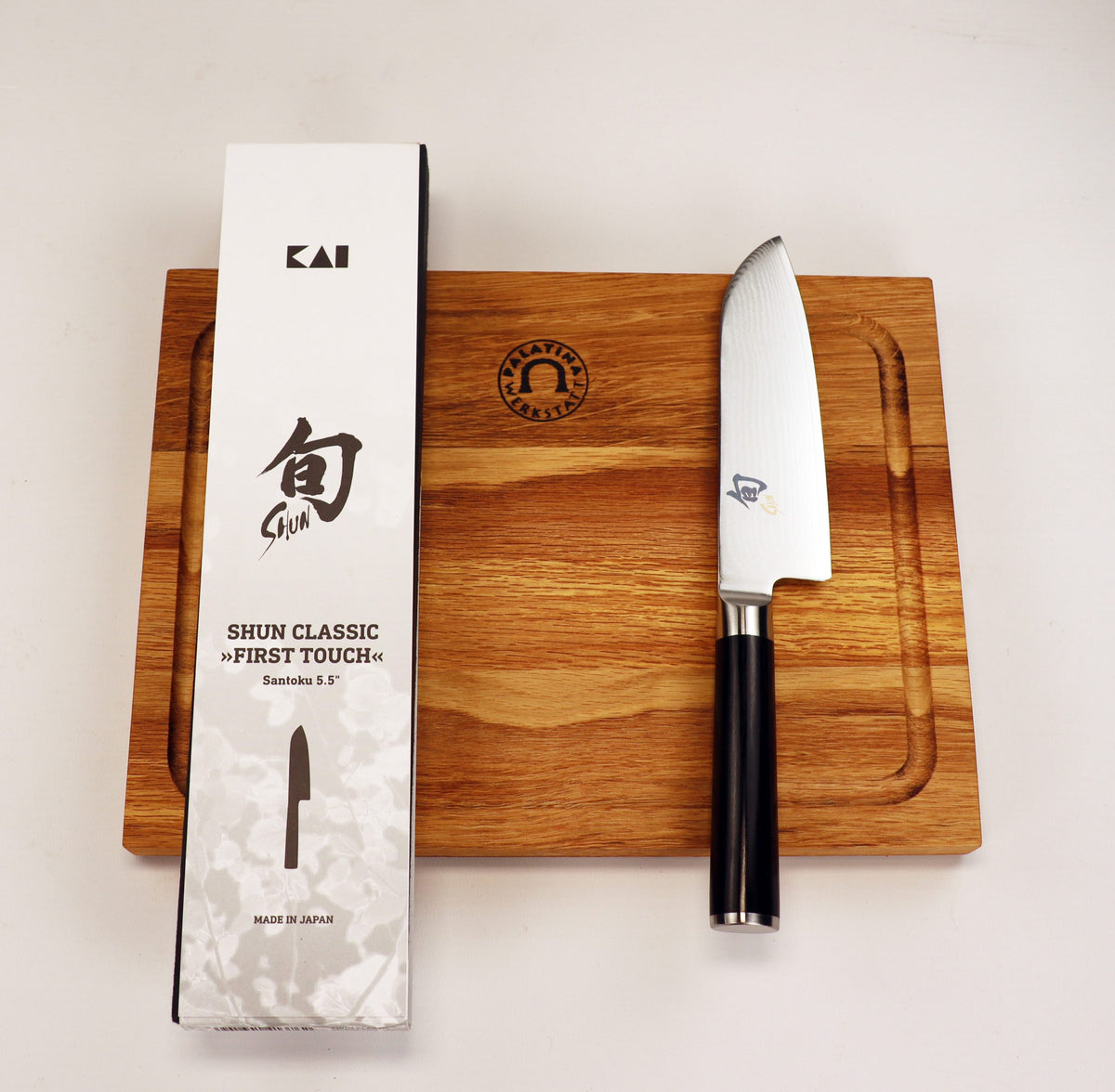 Ein Kai Shun Classic Angebotsset DM-0727, bestehend aus einem ultrascharfen 14 cm Santokumesser mit schwarzem Griff und einem massiven Schneidebrett aus Eiche (30x18 cm), liegt samt der japanisch beschrifteten Box bereit.