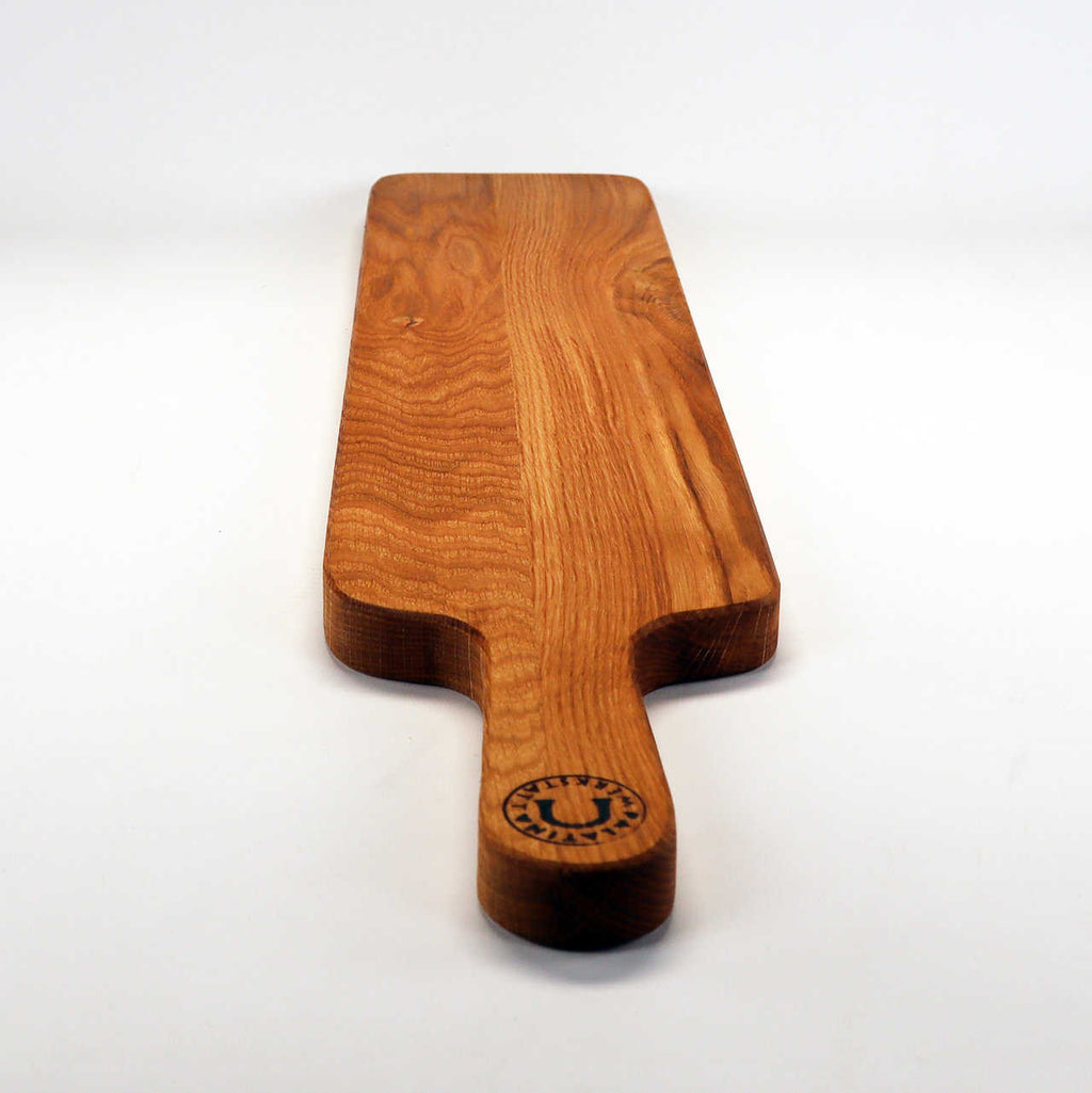 Ein rechteckiges Holzschneidebrett mit Griff, gefertigt aus altem Weinfassholz. Die Oberfläche weist eine natürliche Holzmaserung auf und der Griff ist am Ende mit einem runden schwarzen Logo versehen. Perfekt als Käsebrett vor einem schlichten weißen Hintergrund. Dieses exquisite Stück ist bekannt als Palatina Werkstatt | handgefertigtes Baguettebrett, Käsebrett aus altem Weinfassholz.