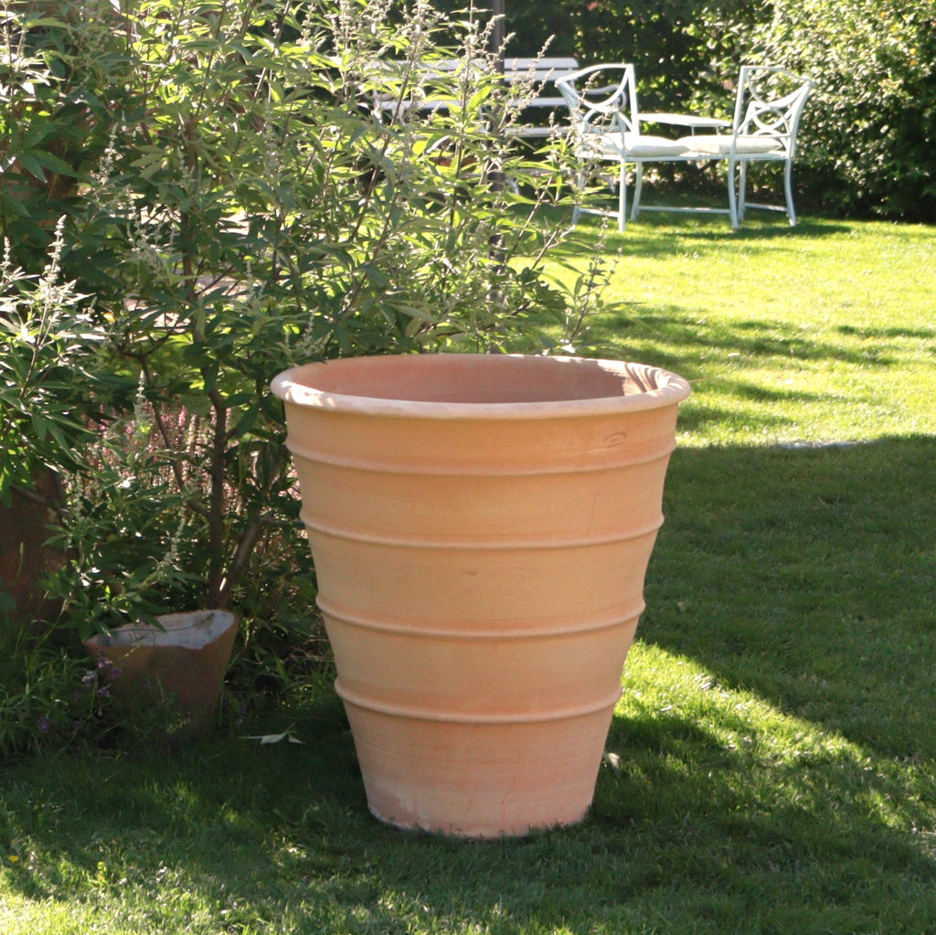 Ein großer, leerer Blumenkübel hoch „Yucca“ (H=80 cm) aus Terrakotta steht auf einer Rasenfläche. Im Hintergrund stehen ein weißer Metalltisch und Stühle im Schatten hoher Pflanzen und Bäume und schaffen eine friedliche Gartenatmosphäre.