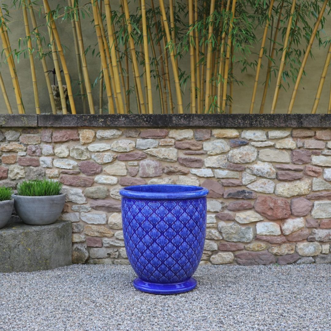Der Glasierte Pflanzkübel "Indigo Provence" in Kobaltblau (Höhe 30,5-54 cm) steht auf Kies an einer Steinmauer, daneben stehen ein hoher Bambus und zwei kleine graue Grasblumentöpfe.
