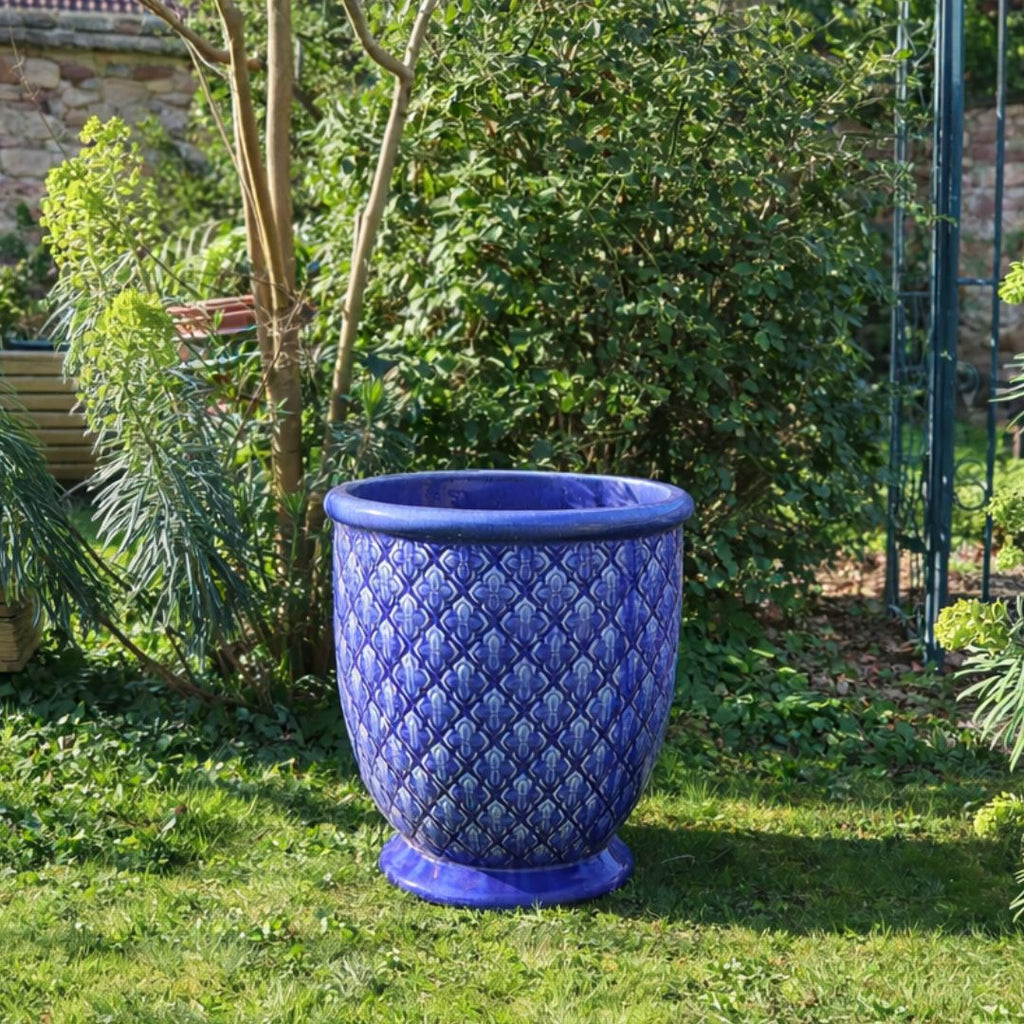 Ein großer, verschnörkelter glasierter Pflanzkübel "Indigo Provence" in Kobaltblau (Höhe 30,5-54 cm) mit Muster steht leer auf einer Wiese, umgeben von grünen Büschen und Bäumen in einem sonnigen Garten.