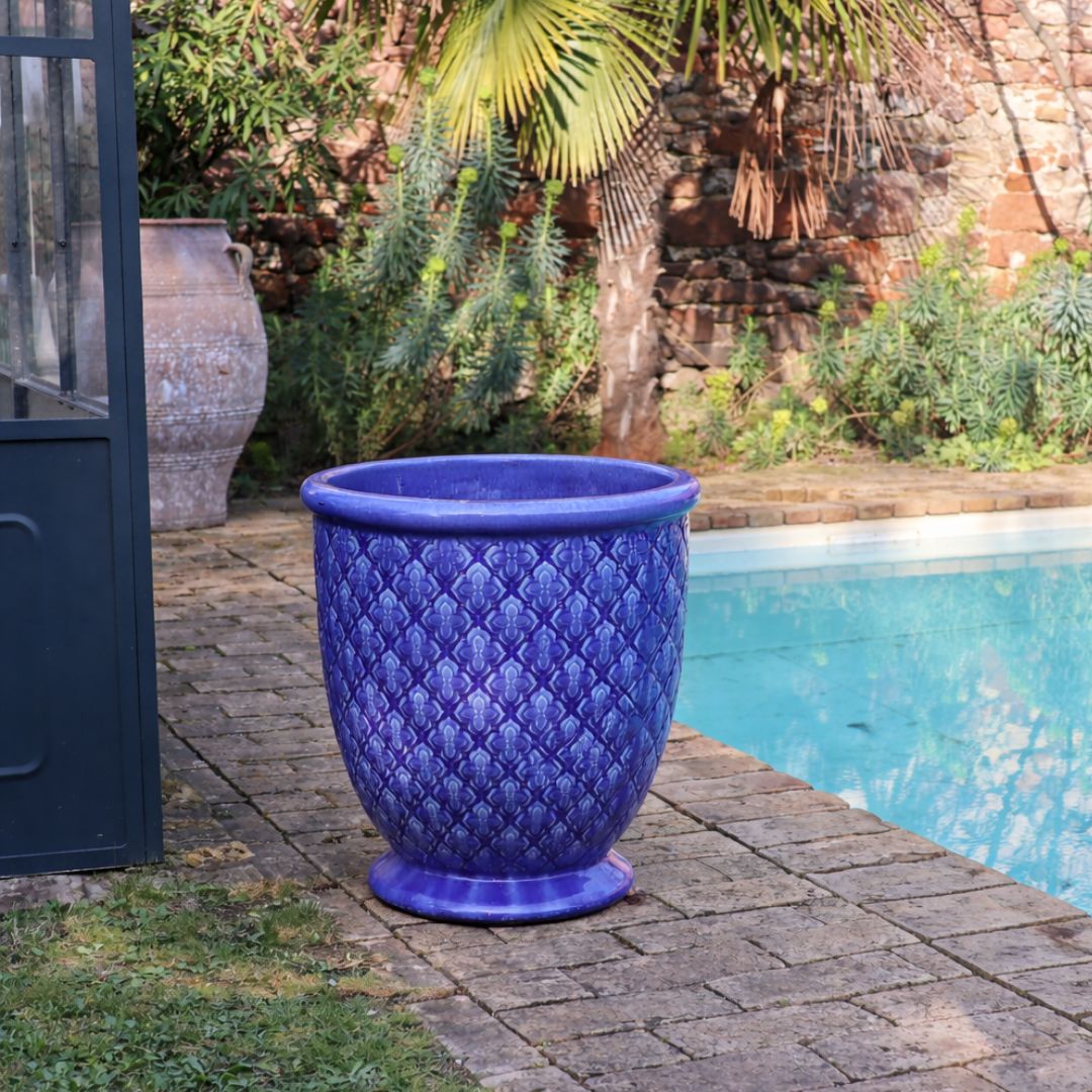 Der glasierte Pflanzkübel "Indigo Provence" in Kobaltblau (Höhe 30,5-54 cm) steht auf Steinfliesen neben einem Pool, umgeben von üppigem Grün und einer hohen Palme im Hintergrund.