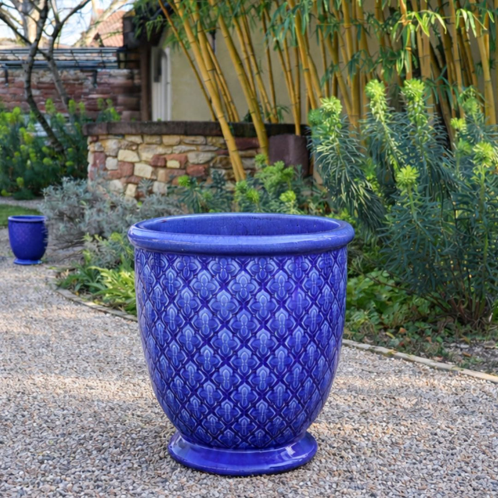 Ein großer, dekorativer blauer Keramiktopf-Glasierter Pflanzkübel "Indigo Provence" in Kobaltblau (Höhe 30,5 bis 54 cm)- steht auf einem Kiesweg im Garten, umgeben von Grünpflanzen und Bambus.
