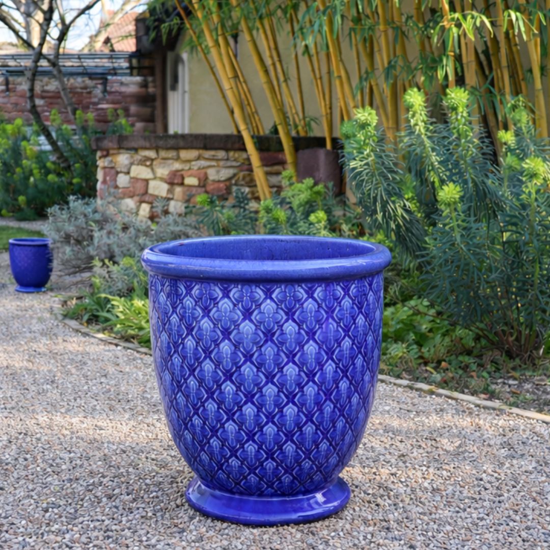 Ein großer, dekorativer blauer Keramiktopf-Glasierter Pflanzkübel "Indigo Provence" in Kobaltblau (Höhe 30,5 bis 54 cm)- steht auf einem Kiesweg im Garten, umgeben von Grünpflanzen und Bambus.