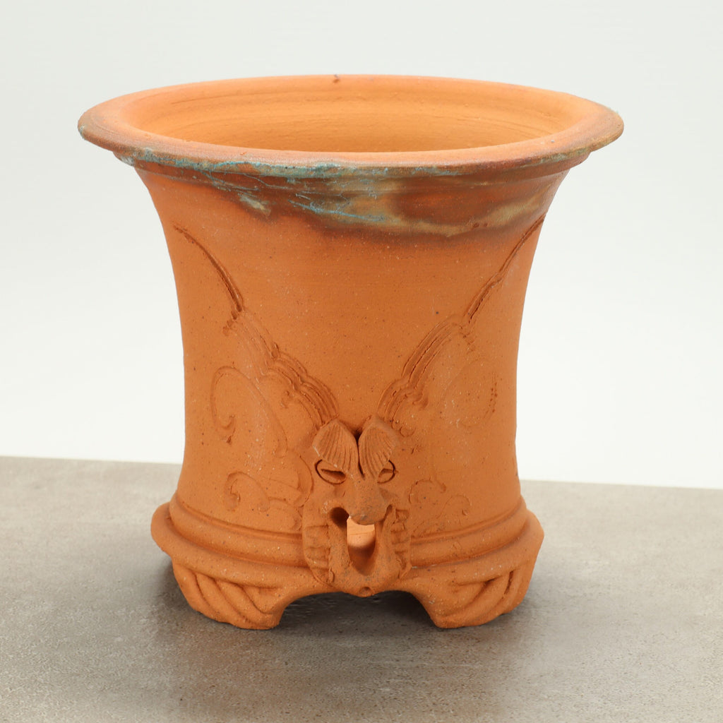 Ein dekorativer Terrakotta-Keramiktopf mit breitem, ausgestelltem Rand. Der handgefertigte Terrakotta-Blumentopf weist am Boden ein strukturiertes Design mit einem Drachengesicht auf und hat einige grünlich-blaue Patinaflecken am Rand. Er steht auf einer flachen, hellgrauen Oberfläche vor einem schlichten Hintergrund.