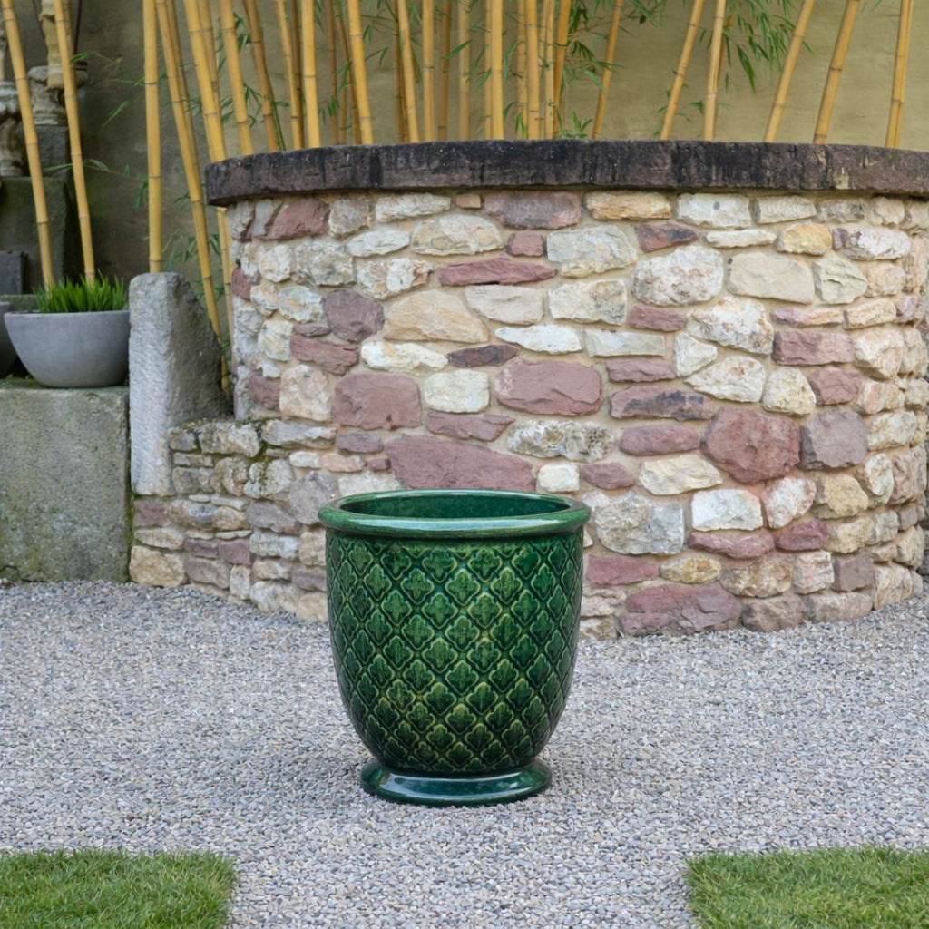 Ein großer grüner Keramikübertopf mit Rautenmuster steht auf Kies vor einem runden Steinbrunnen, mit Bambuspflanzen und Grünzeug im Hintergrund.