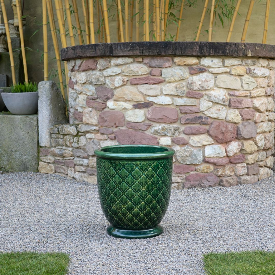 Ein großer grüner Keramikübertopf mit Rautenmuster steht auf Kies vor einem runden Steinbrunnen, mit Bambuspflanzen und Grünzeug im Hintergrund.