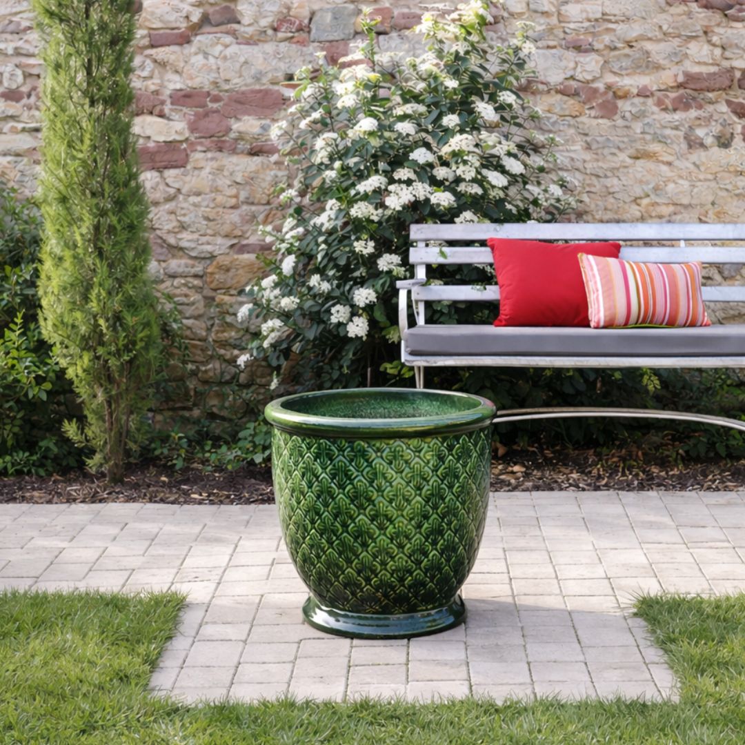 Ein grünes Keramikgefäß steht auf einer gepflasterten Terrasse in einem Garten mit Gras, einem blühenden Strauch, einer Steinmauer und einer weißen Bank mit roten und gestreiften Kissen im Hintergrund.