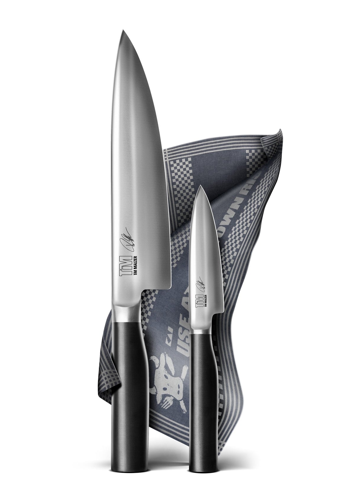 Zwei schlichte Tim Mälzer Kamagata Küchenmesser, jeweils mit schwarzem Griff, werden elegant in der KAI X-MAS SELECTION DREAMBOX präsentiert. Ein Messer ist größer und das andere kleiner, beide stehen aufrecht vor einem teilweise gefalteten dunklen Tuch mit Karomuster und grafischem Design und bilden so ein stilvolles Display.