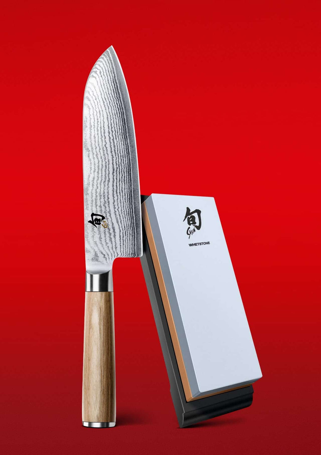 Eine KAI X-MAS SELECTION DREAMBOX mit dem Shun Classic White Santokumesser mit Holzgriff steht elegant aufrecht neben einem Schleifstein. Der leuchtend rote Hintergrund hebt das charakteristische Damaszenerstahlmuster auf der Klinge des Messers hervor.