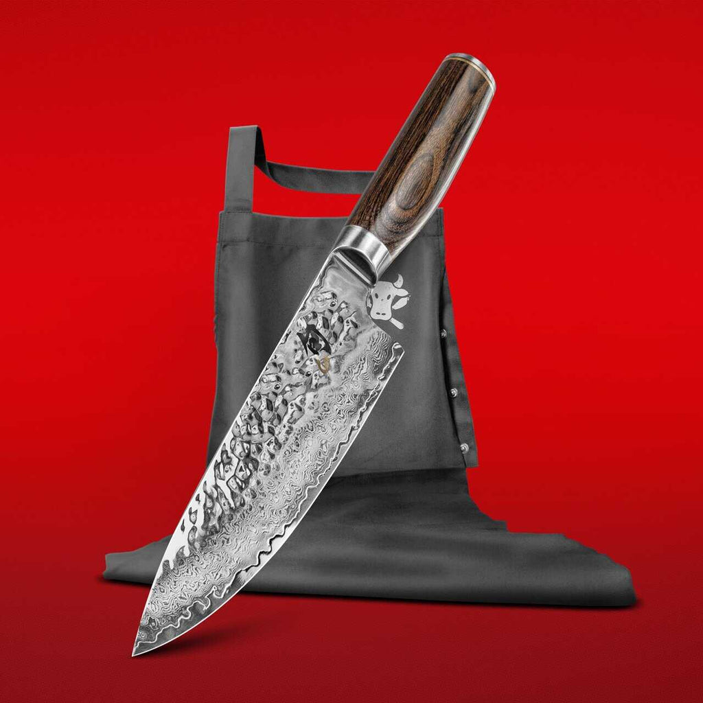 Die KAI X-MAS SELECTION DREAMBOX enthält ein Kochmesser mit kunstvoll gemusterter Klinge und elegantem Holzgriff, der nach unten zeigt. Es lehnt an einer gefalteten schwarzen Tim Mälzer Kochschürze, auf der ein kleines, stilisiertes Cartoon-Kochsymbol abgebildet ist. Der Hintergrund ist in leuchtendem Rot gehalten.