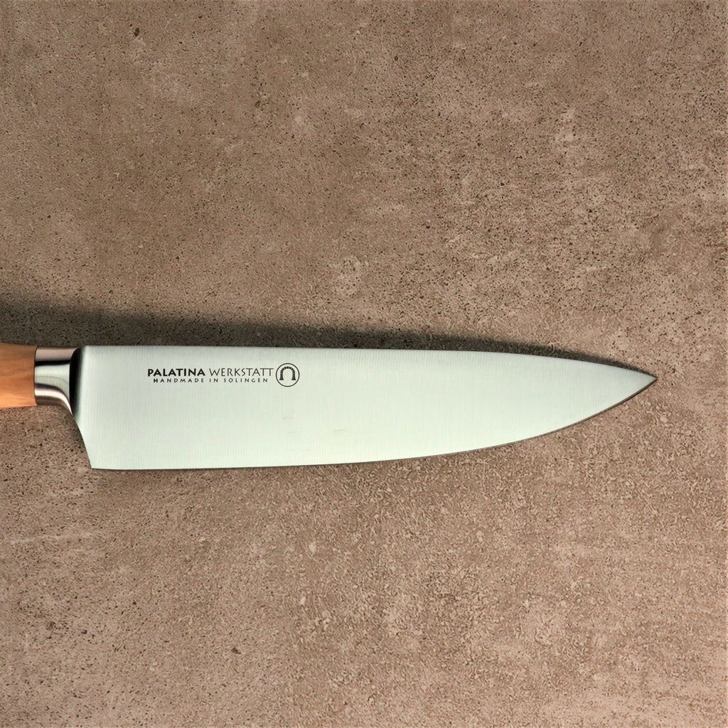 Das Kochmesser der Palatina Werkstatt Messerserie mit 21 cm langer Klinge und Olivenholzgriff liegt flach auf einer strukturierten, grauen Oberfläche. Die Klinge trägt die Inschrift „PALATINA WERKSTATT Handmade in Solingen“, was auf die handwerkliche Qualität hinweist. Das Kochmesser liegt mit der Schneide nach oben und dem Griff nach links.