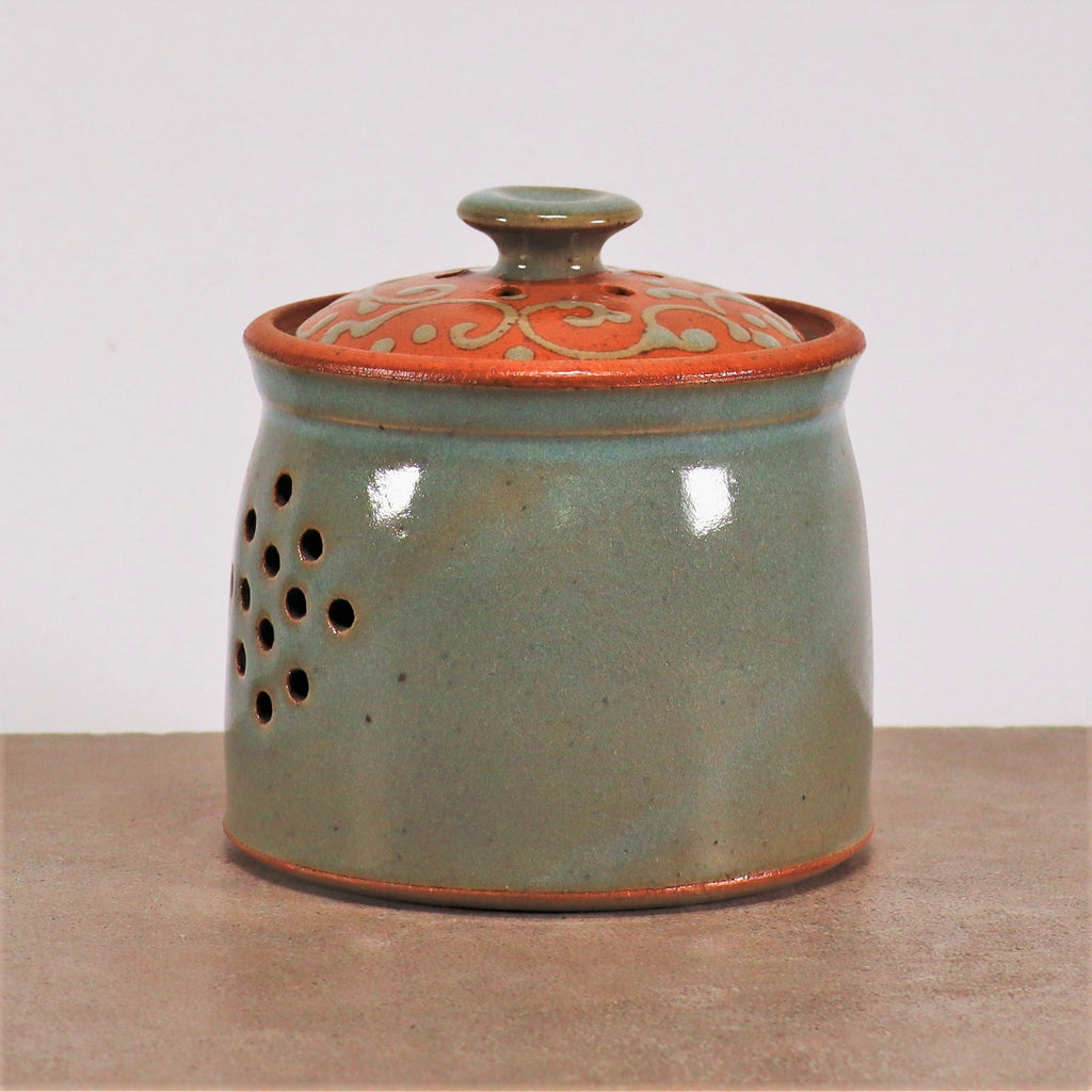 Ein glasierter Zwiebel- und Kartoffeltopf, handgefertigt aus Steinzeug, mit einer hellgrünen Glasur und dekorativen Löchern an der Seite. Der Deckel hat eine metallisch orange Farbe mit komplizierten Wirbelmustern am Rand. Der runde Steinzeugbehälter steht auf einer schlichten Oberfläche vor einem weißen Hintergrund.