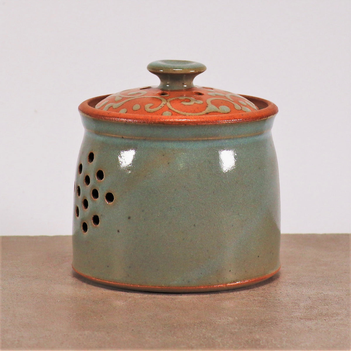 Ein glasierter Zwiebel- und Kartoffeltopf, handgefertigt aus Steinzeug, mit einer hellgrünen Glasur und dekorativen Löchern an der Seite. Der Deckel hat eine metallisch orange Farbe mit komplizierten Wirbelmustern am Rand. Der runde Steinzeugbehälter steht auf einer schlichten Oberfläche vor einem weißen Hintergrund.