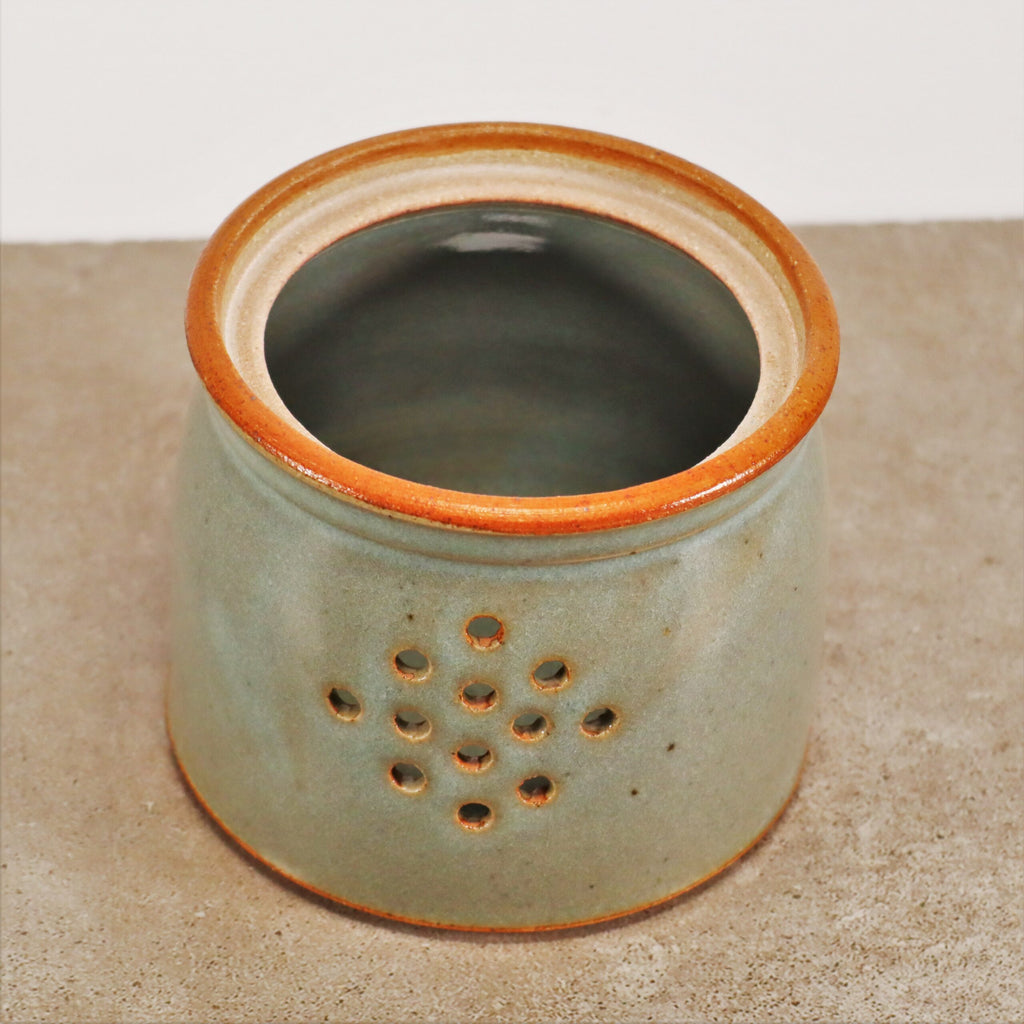 Ein kleiner, zylindrischer Keramikbehälter mit grünlicher Glasur und braun umrandeter Öffnung. Der Glasierte Zwiebel- und Kartoffeltopf, handgefertigt aus Steinzeug im traditionellen Stil, weist auf einer Seite eine Reihe kleiner Löcher auf, die in einem Dreiecksmuster angeordnet sind und zur Belüftung oder zum Durchlassen von Licht dienen.