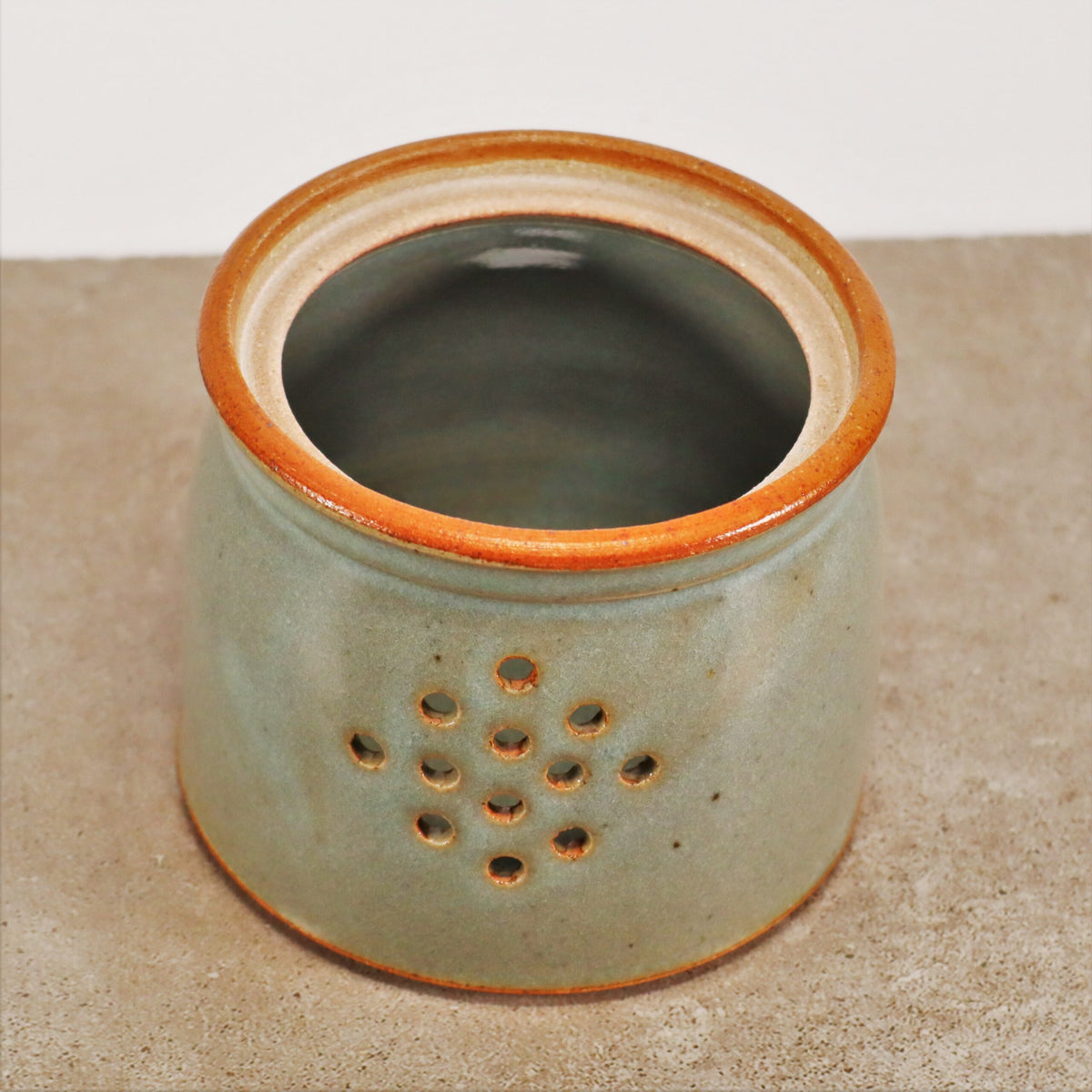 Ein kleiner, zylindrischer Keramikbehälter mit grünlicher Glasur und braun umrandeter Öffnung. Der Glasierte Zwiebel- und Kartoffeltopf, handgefertigt aus Steinzeug im traditionellen Stil, weist auf einer Seite eine Reihe kleiner Löcher auf, die in einem Dreiecksmuster angeordnet sind und zur Belüftung oder zum Durchlassen von Licht dienen.