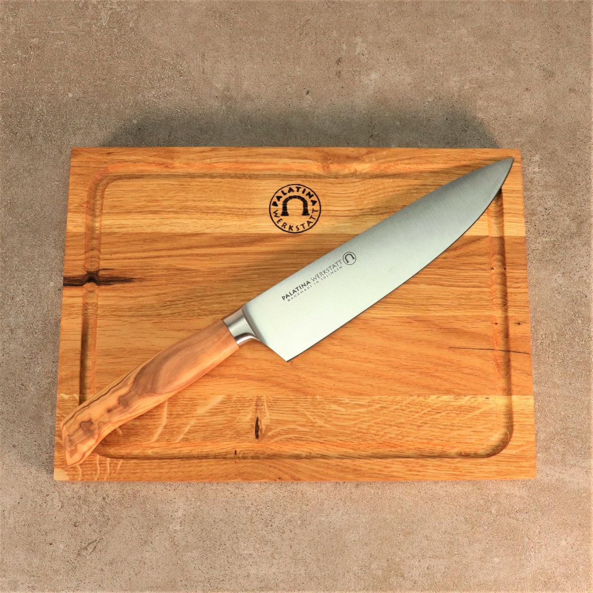 Ein scharfes Palatina Werkstatt Messerserie Kochmesser mit 21 cm Klinge und Olivenholzgriff liegt auf einem hölzernen Schneidebrett. Die Klinge des Messers trägt die Aufschrift „Palatina Messer“ und ein Logo. Das Schneidebrett ist rechteckig mit einer Rille um den Rand und einem Logo oben in der Mitte.