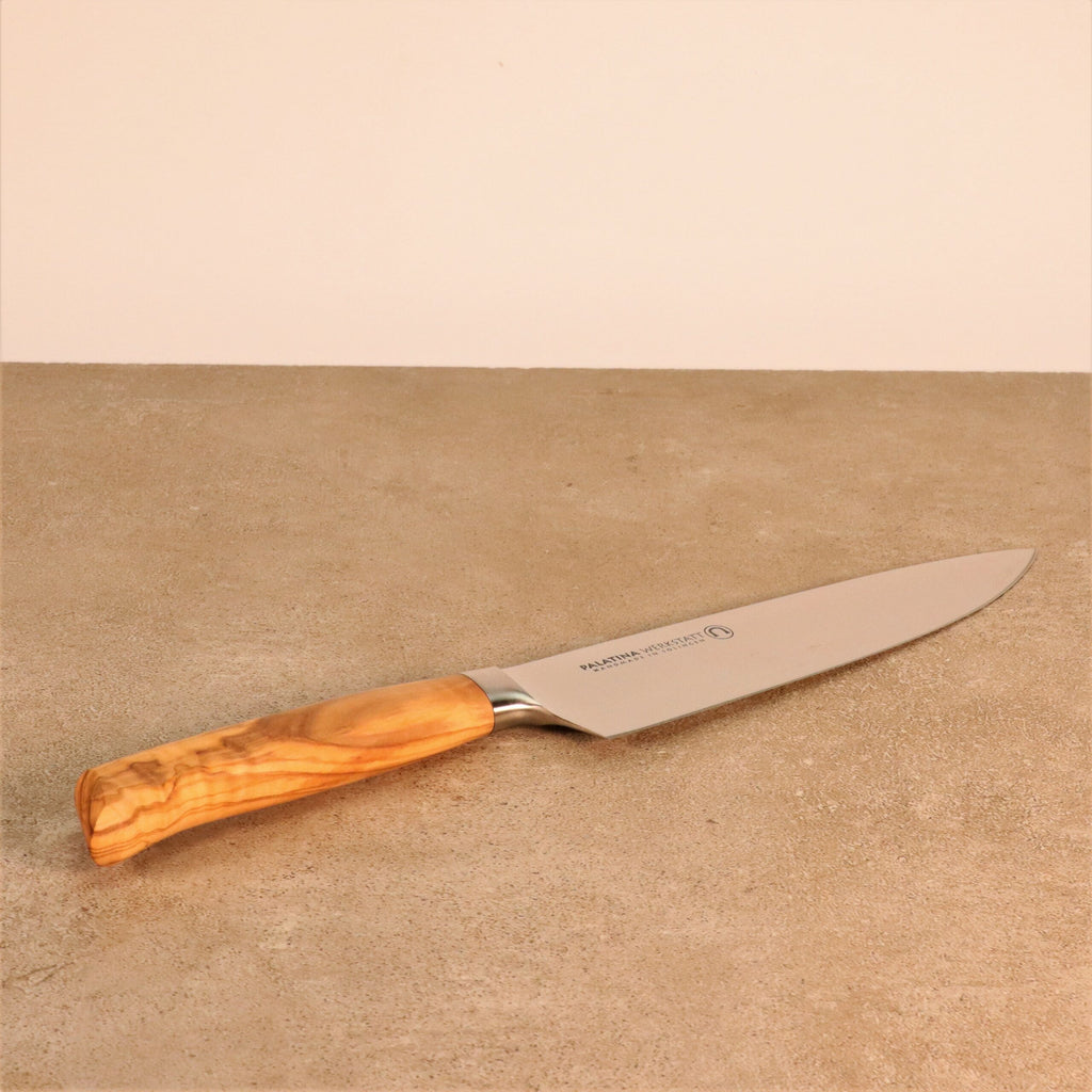 Ein hochwertiges Kochmesser der Palatina Werkstatt Messerserie mit 21 cm langer Klinge und Olivenholzgriff liegt auf einer beigen Steinplatte. Die Klinge ist glänzend und scharf, und der Griff hat eine glatte Oberfläche, die die natürliche Holzmaserung zur Geltung bringt. Das gut verarbeitete Messer der Palatina Werkstatt liegt mit der Klinge nach rechts zeigend.