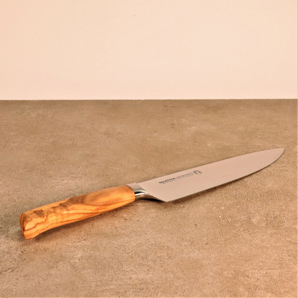 Ein scharfes Küchenmesser mit einem Griff aus alten Fassdauben und Olivenholzgriff liegt auf einer beigen Arbeitsplatte vor einer hellen Wand. Die Klinge besteht aus rostfreiem Stahl und der Griff weist eine natürliche Holzmaserung auf.
